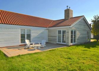An L-shaped, wood-panelled Essex holiday bungalow with a wooden deck containing a table and sunlounger all overlooking a large lawn.