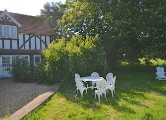 Outdoor dining chairs cluster around a table on a tree-lined lawn on Osea Island. Behind them is a cross-timbered holiday cottage on Osea Island.