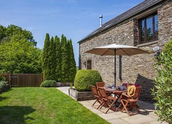 A table with parasol and chairs tands on a sunny patio behind a stone built holiday cottage in Devon.