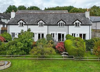 A terrace of 4 slate-roofed cottages over looking a lawn with tall shrubs and an open field in Dittisham. 