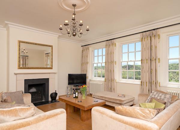 A stylish lounge with superb rural views through its triple windows. It is furnished with two large sofas with svatter cushions, a coffee table, TV and  an open fire place.