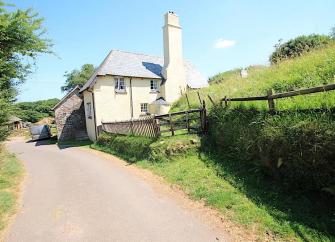 L-shaped exterior of a country cottage with a lawn surrounded by flowering shrubs and bushes.