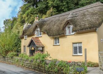 Exterior of a thatched cottage with porch and low stone wall.