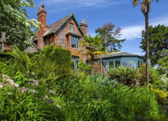 A House with large conservatory is surrounded by a garden with lawns and mature shrubs.