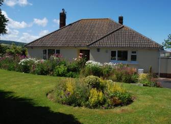 A bungalow sits behind a large lawn with flower-filled borders.