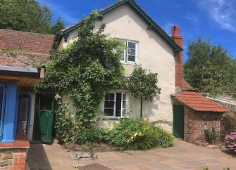 The gable end of a 2-storey wisteria-clad Dunster Cottage with a paved courtyard in front.
