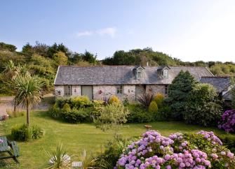 A stone cottage overlooks a lawn bordered by flowering shrubs.