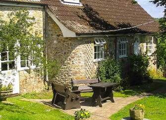 A stone-built holiday cottage with a picnic table patio overlooks a lawn.
