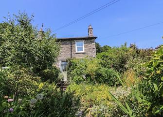 A stone-builkt cottage overlooks a garden with mature trees and shrubs.