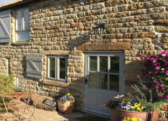 Exterior of a sandstone cottage with a courtyard lined by flowering shrubs.