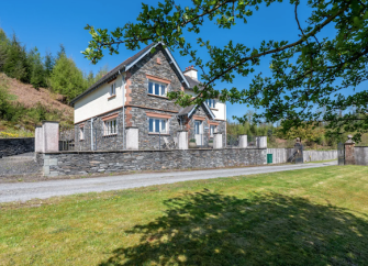 A Large stone-built Lake District lodge overlook a drive and large lawn.