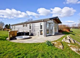 A wooden eco-lodge with a hot tub stands in an open field.