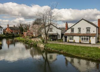 A Herefordshire riverside cottage with bay windows is fronted by a grassy bank down to a wide river.