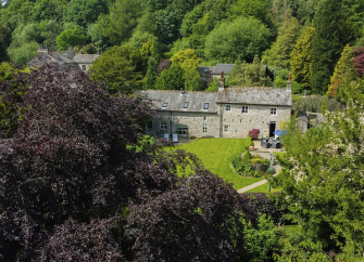 A stone-built Lake District Cottage sits in a woodland clearing surrounded by mature trees.