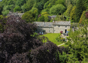 A stone-built LAke District Cottage sits in a woodland clearing surrounded by mature trees.