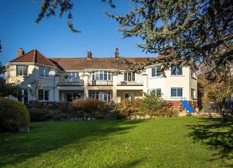 A large house with two bay wings overlooks a shrub-liined lawn.