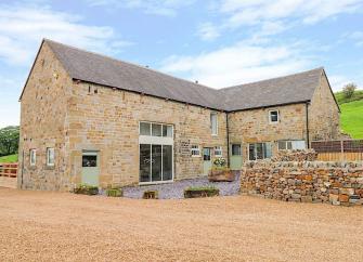 A large, stone-built contemporary cottage with a stone wall, surrounded by countryside.