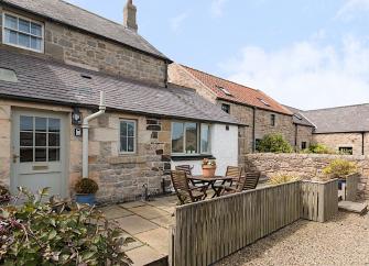 Exterior of a stone-built holiday cottage with ground fllor extension behind a paveed  garden with dining table and chairs.