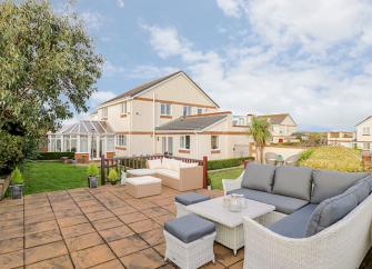 A modern, detached Anglesey holiday home surrounded by lawns and paved terraces with seating.