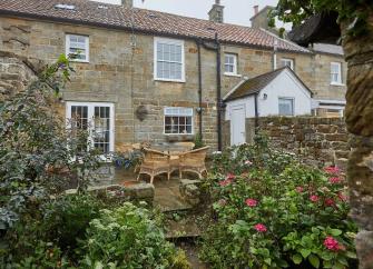 Exterior of a stone built holiday cottage in Whitby with a flagstoned front garden containing outdoor dining facilities.