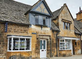 A semi-detached Cotswold holiday cottage with leaded, bay windows overlooks a village street.