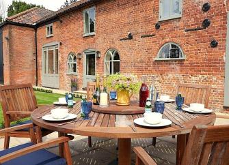 A wood dining table set for breakfast stands on a courtyard in front of a brick-built Norfolk cottage with arched windows.