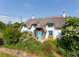 A thatched Hampshire holiday cottage sits behind a flowering hedgerow in the countryside.