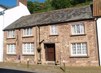 Exterior of a double-fronted stone holiday cottage in Dunster nestling into a steep wooded hillside.