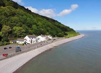 A thatched cott`ge lings to the foot of a steeply wooded hill right above a pebble beach.