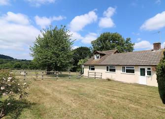 A single-storey Dorset holiday cottage overlooks a large lawn surrounded by a wooden fence, trees and rose bushes.