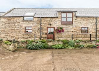 Exterior ofva semi-detached Alnwick barn conversion overlooking a small flowerbed and paved courtyard.