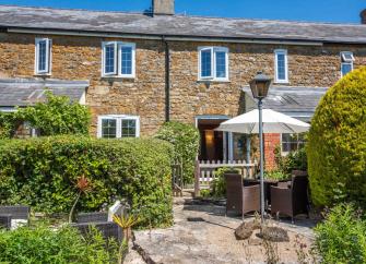 Exterior and front garden of a stone-built Dorset holiday cottage with outdoor dining furniture on its terrace.