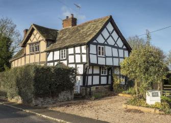 A half-timbered Tudor cottage  in Leominster overlooks a shingle parking space and shrub-filled garden.