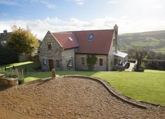 L-shaped exterior of a stone-built cottage surrounded by lawns and a secure hedge.