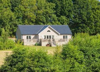 A modern single storey lodge stands in front of trees overlooking a hedge-lined field.