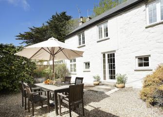 A stone-built cottage overlooks a shrub-lined terrace with dining table chairs and a parasol