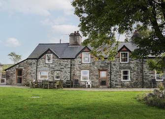 A wide, 2-storey, stone house in Snowdonia with three gable ends flanked by single storey wings facing a large, level lawn with mature trees.
