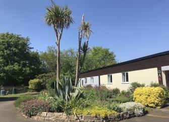 Exterior of a holiday bungalow with a front garden full of shrubs and palm trees.