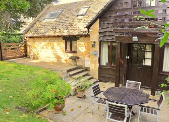 Exterior of a Cotswold honey-stone cottage overlooking a small lawn and patio with a dining table.