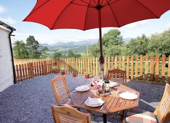 An outdoor dining table underneath a parasol inside a fenced in terrace with rural views beyond. 