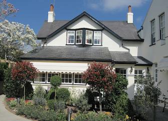 Exterior of a 2-storey cottage with 1st-floor bay windows overlooking a shrub-filled garden.