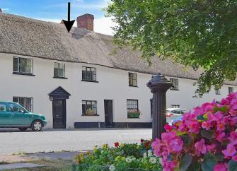 A row of thatched village cottages overlook trees and flowering bushes on a village green.