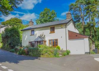 The exterior of a double-fronted Exmoor holiday cottage overlooks a front garden and is surrounded by trees.