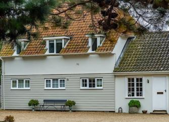 Exterior of a weather-boarded Suffolk Cottage behind a garden lined with fur trees.