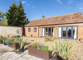 Exterior of a single storey barn conversion with a pair of floor-to-ceiling French doors overlooking a shingled courtyard with flower troughs.