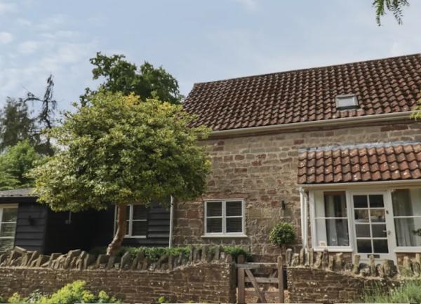 Exterior of  stone-built cottage with a covered porch and front garden with trees behind a stone wall.