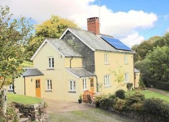 A 2-storey-cottage overlooks a lawn surrounded by trees.
