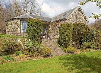 Large single-storey stone-built barn conversion half-hidden by tall shrubs.