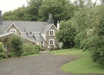 A sweeping drive approaches a twin-gabled Exmoor holiday cottage half-surrounded by mature trees.