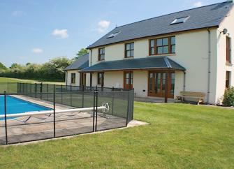 A fenced-in outdoor pool on a lawn overlooked by a large holiday cottage.